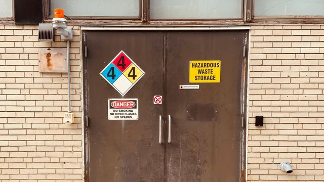 Two metal doors leading to a waste storage area. There are multiple hazard signs on the doors and warnings to not smoke or use open flames nearby. 