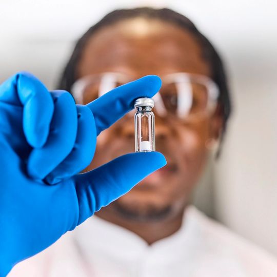 Scientist wearing blue gloves and safety glasses holds up a small vial in a laboratory setting 