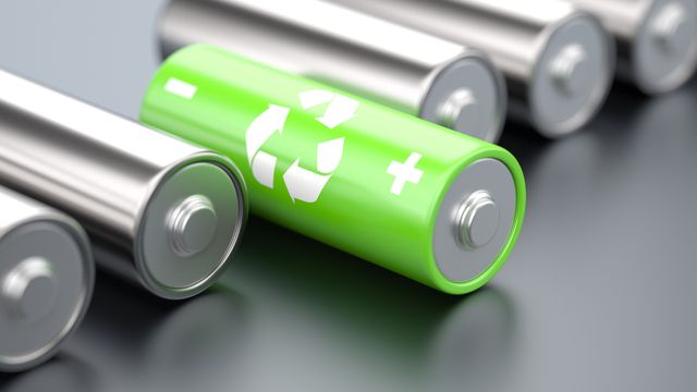 Six AA batteries are laid on a grey table with one of the batteries in the middle in a green colour with a recycling sign 