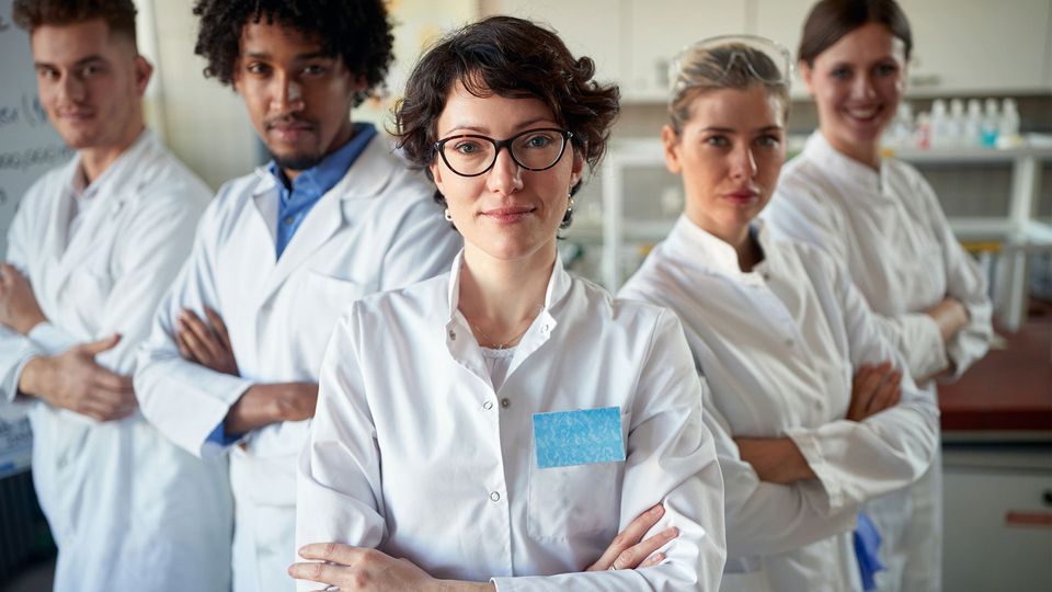 Five scientists in white lab coats standing looking at a camera with their arms folded.